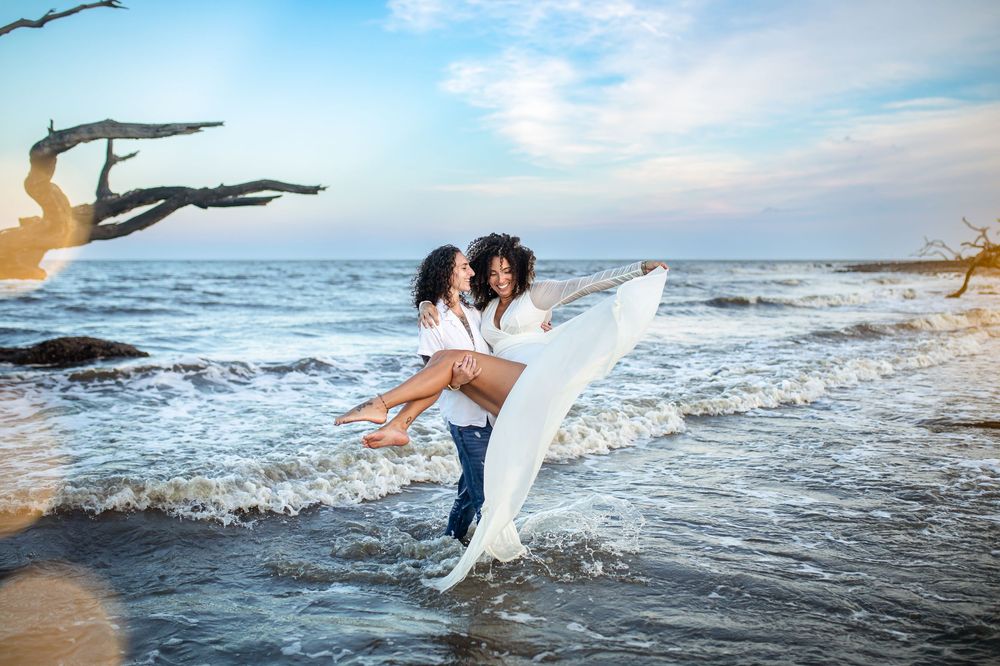Couple embracing on a beach shore, woman in white dress lifted by man in jeans, with ocean waves and driftwood behind them.
