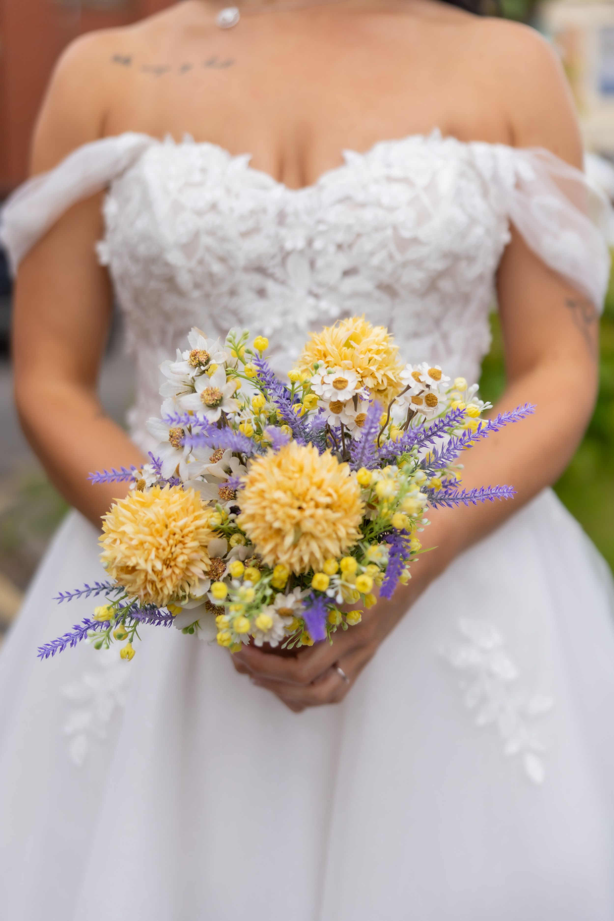 Bride in white off-shoulder lace gown holding a bouquet of yellow dahlias, white daisies, and purple lavender flowers. Casual Captures