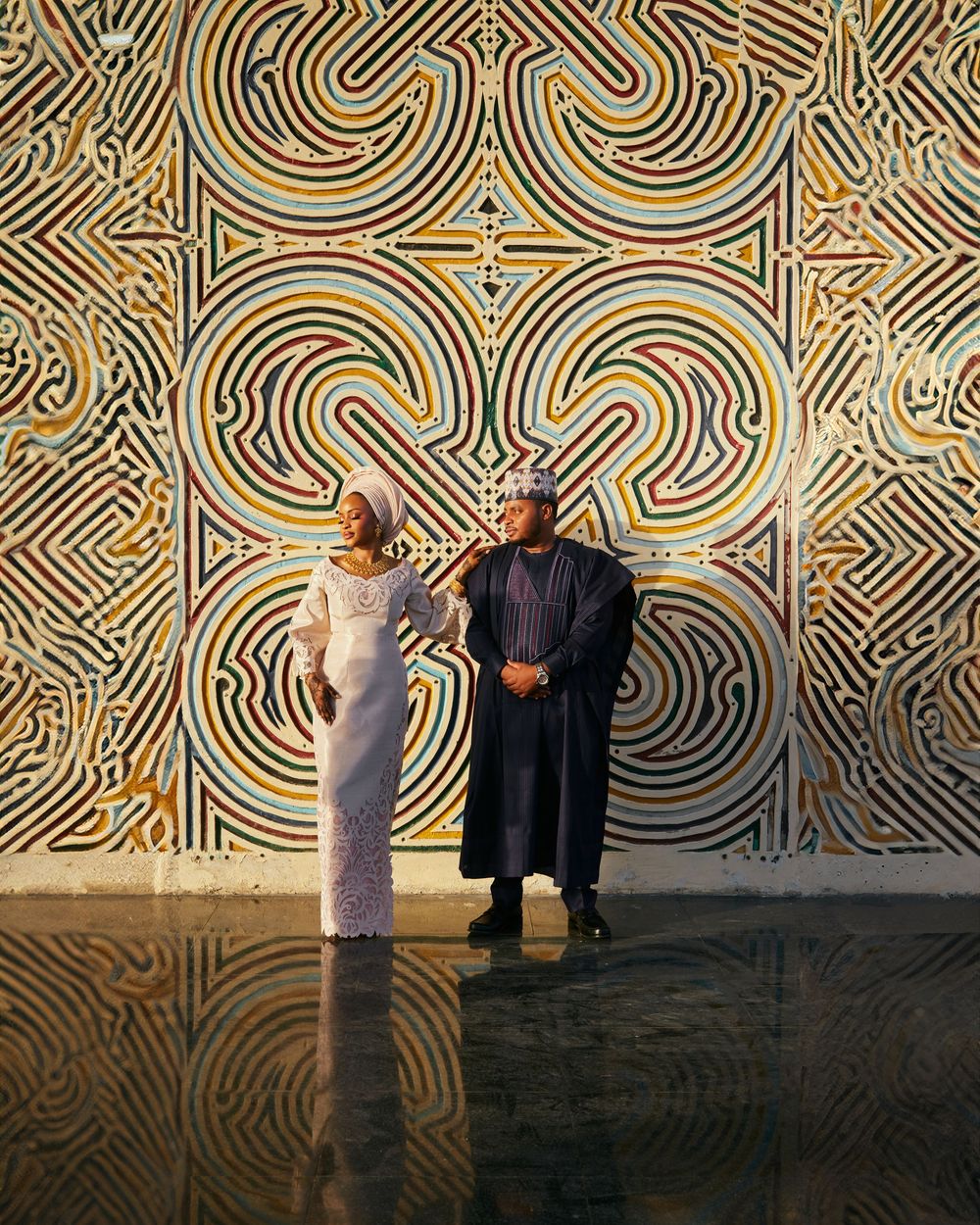 Two figures pose against an ornate gold and black spiral patterned wall in an elegant setting.