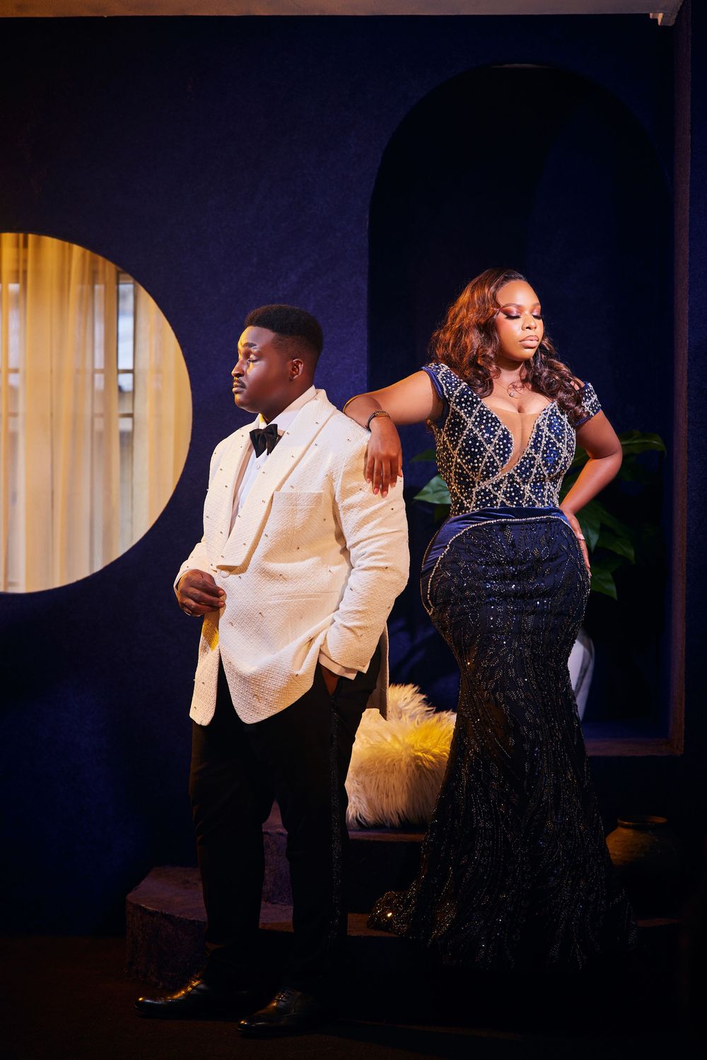 Formal couple in evening wear posing dramatically against a dark blue wall with arched window.