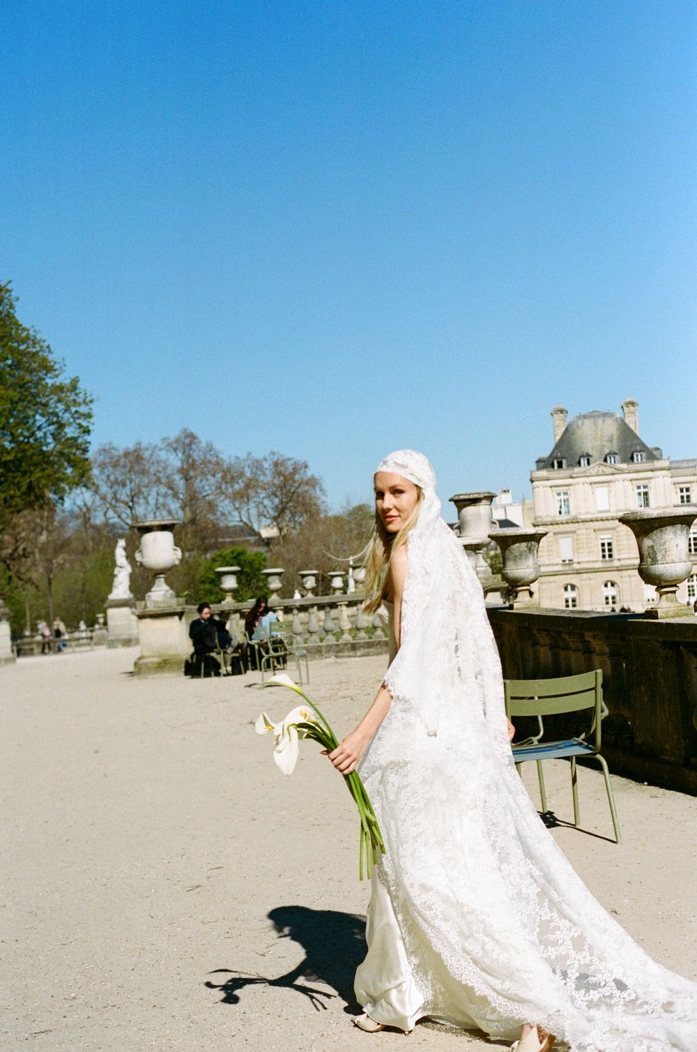 Bride in lace wedding gown holding white calla lilies poses in sunlit Jardin du Luxembourg, Paris.