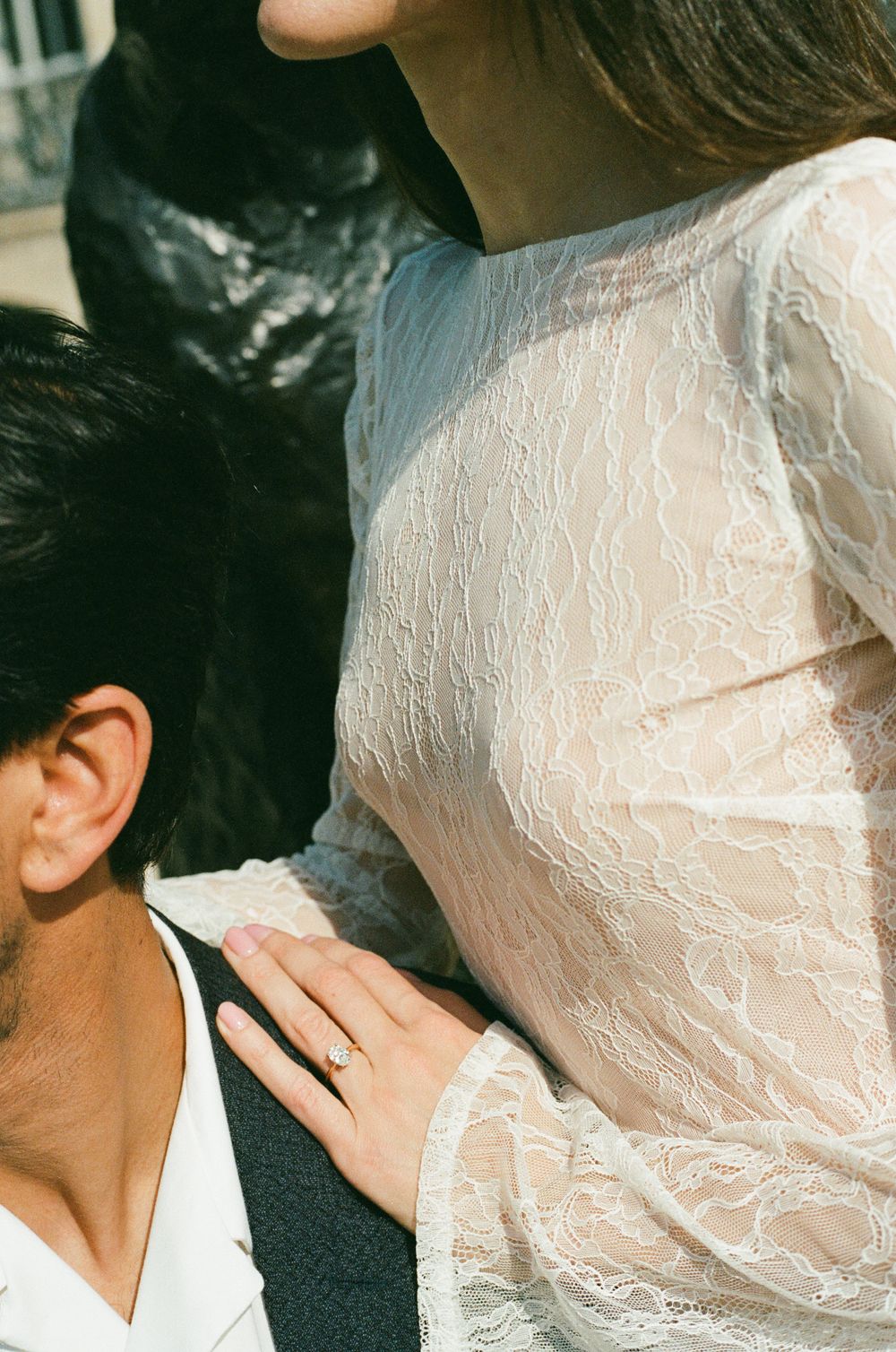 A woman in a sheer white lace dress places her hand on a man's shoulder, showcasing an engagement ring.