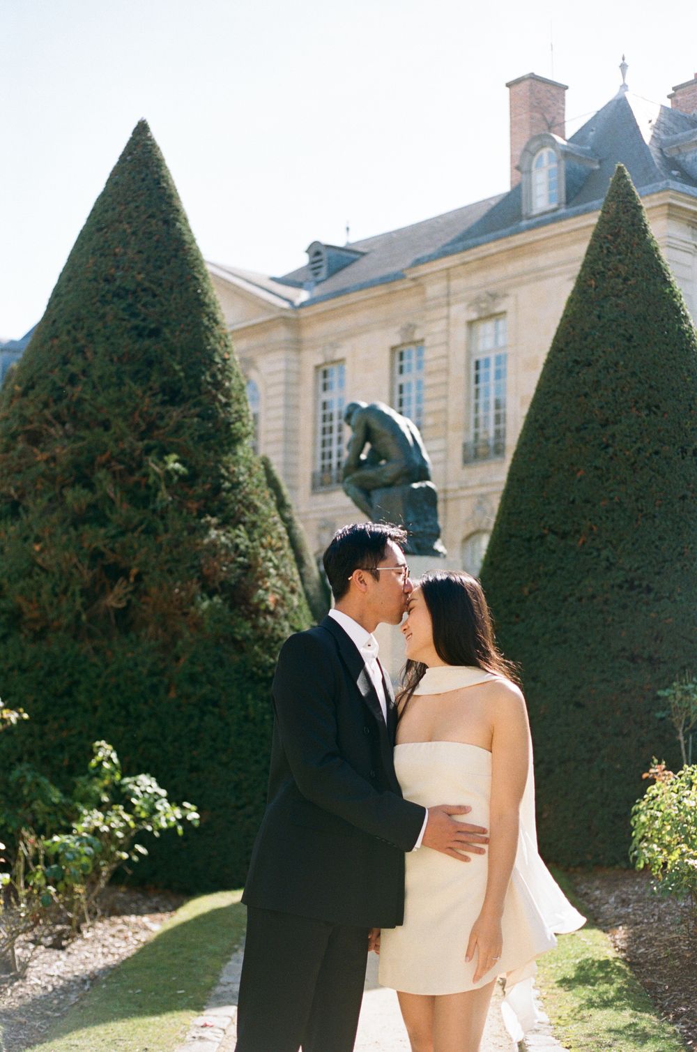 Couple sharing a romantic moment in a French garden with The Thinker sculpture and elegant mansion backdrop.
