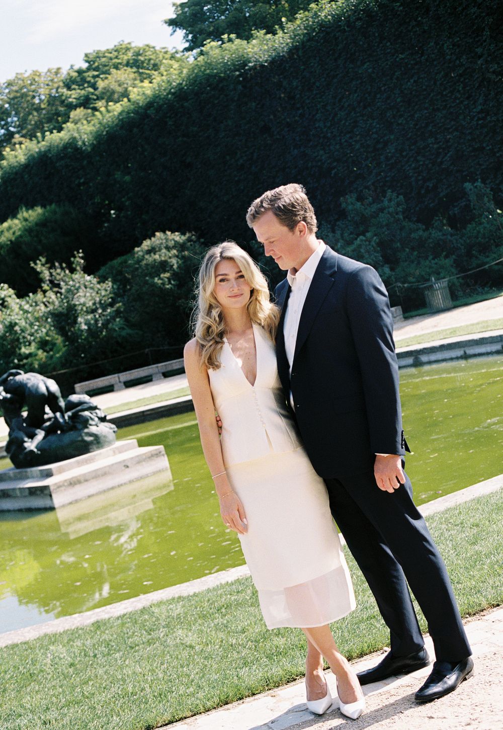Elegant couple posing in formal attire by a garden pond, woman in white midi dress, man in dark suit.