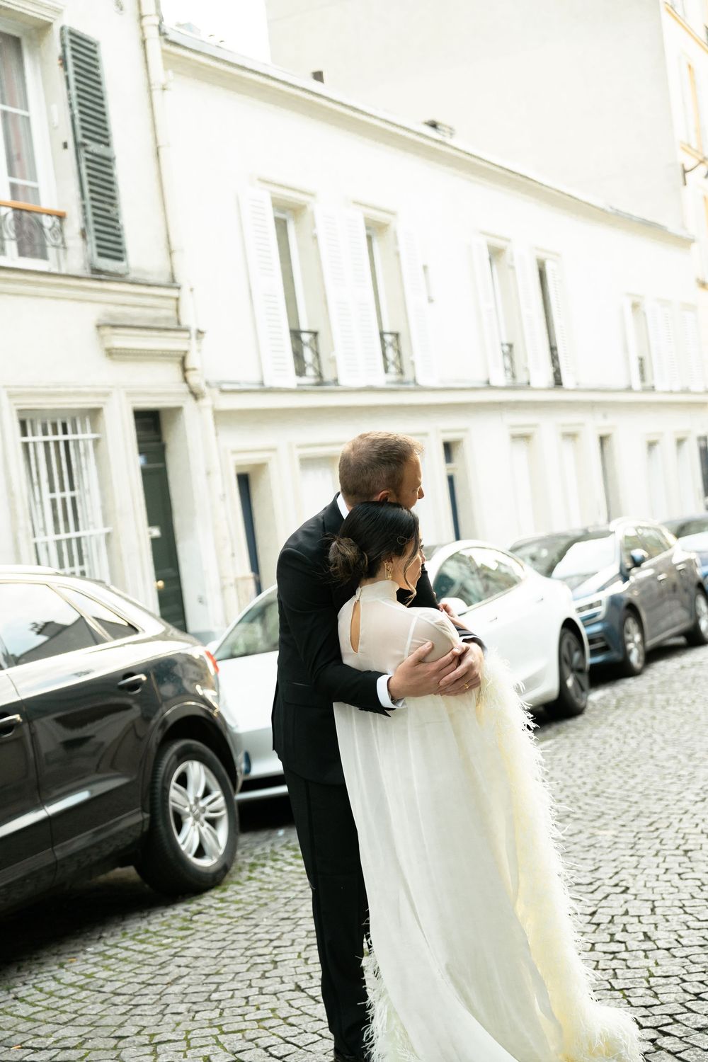 Bride and groom embracing on a cobblestone Paris street, she in a flowing white gown, he in a black suit.