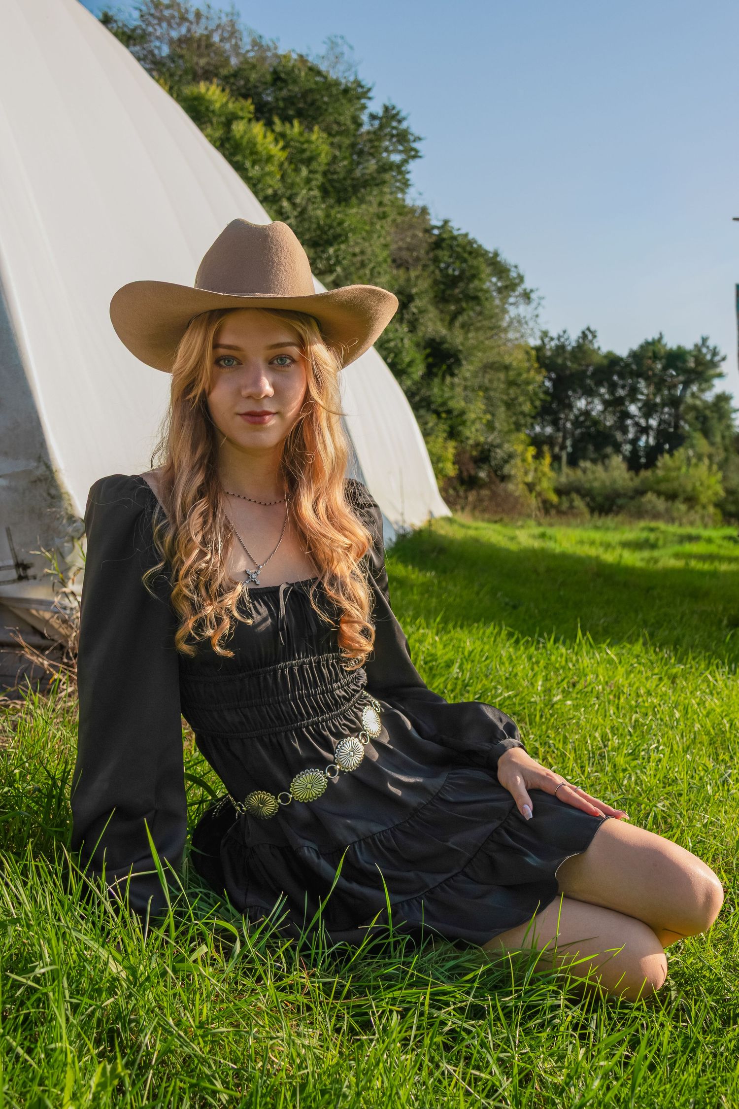 Beautiful blonde girl sitting in the sun. Western styled shoot. Black dress. Cowboy hat. Tall green grass. Farm Yard
