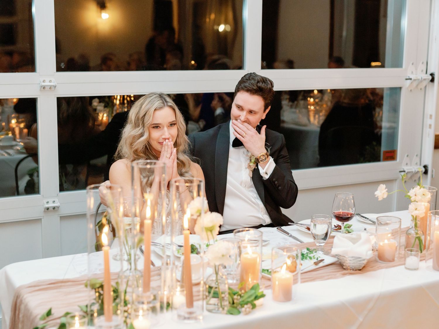 A bride and groom sit at the head table of their reception with teary eyes and hands covering their mouths.
