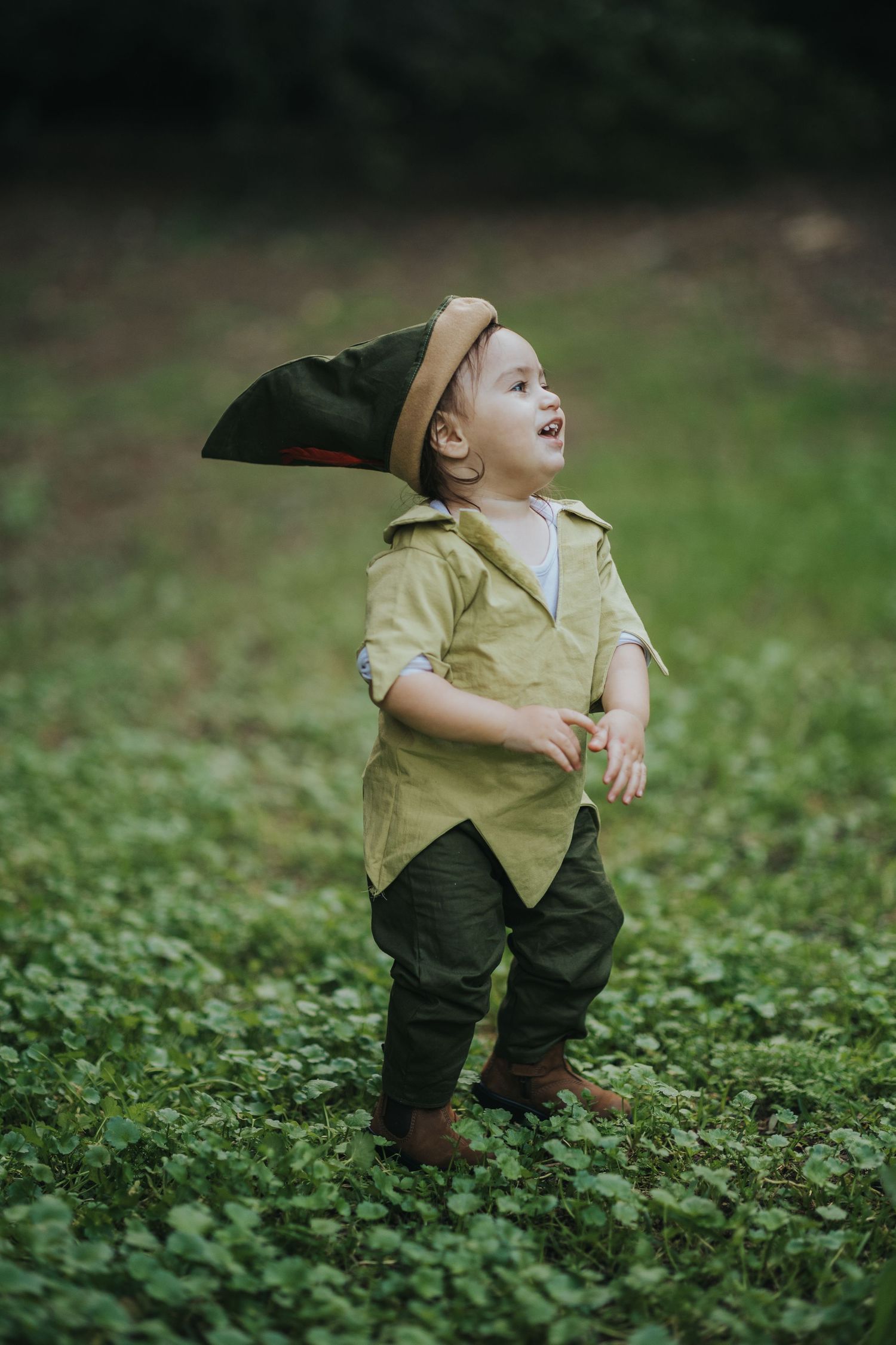 A small child in a classic Peter Pan inspired costume plays joyfully in a field of green clovers.