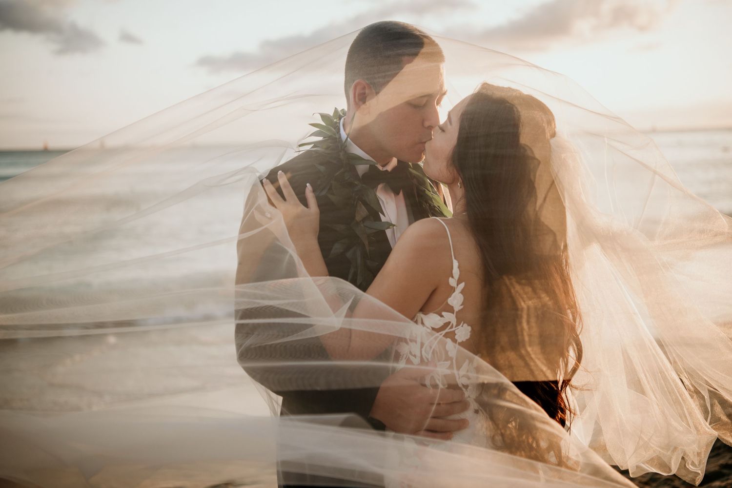 A romantic couple embraces on a beach at sunset with a flowing veil creating an ethereal atmosphere.