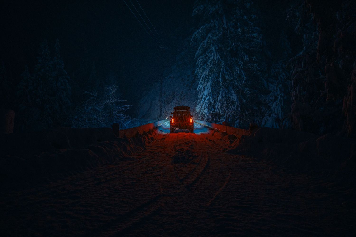 Red car illuminated in dark snowy forest at night with glowing headlights casting warm light on snow-covered ground.