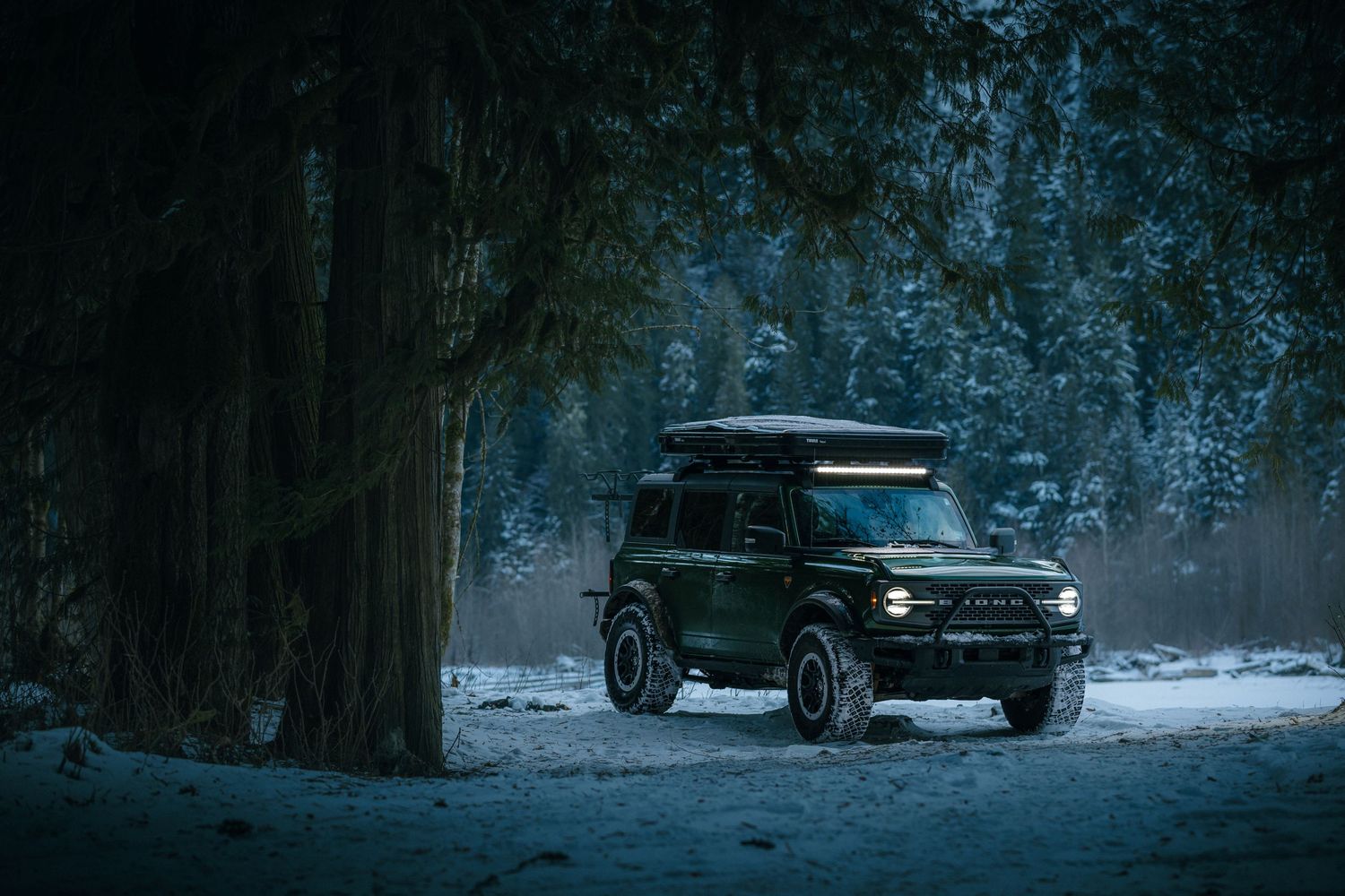 A black SUV parked on a snow-covered forest road at dusk with moody blue lighting and atmospheric surroundings.