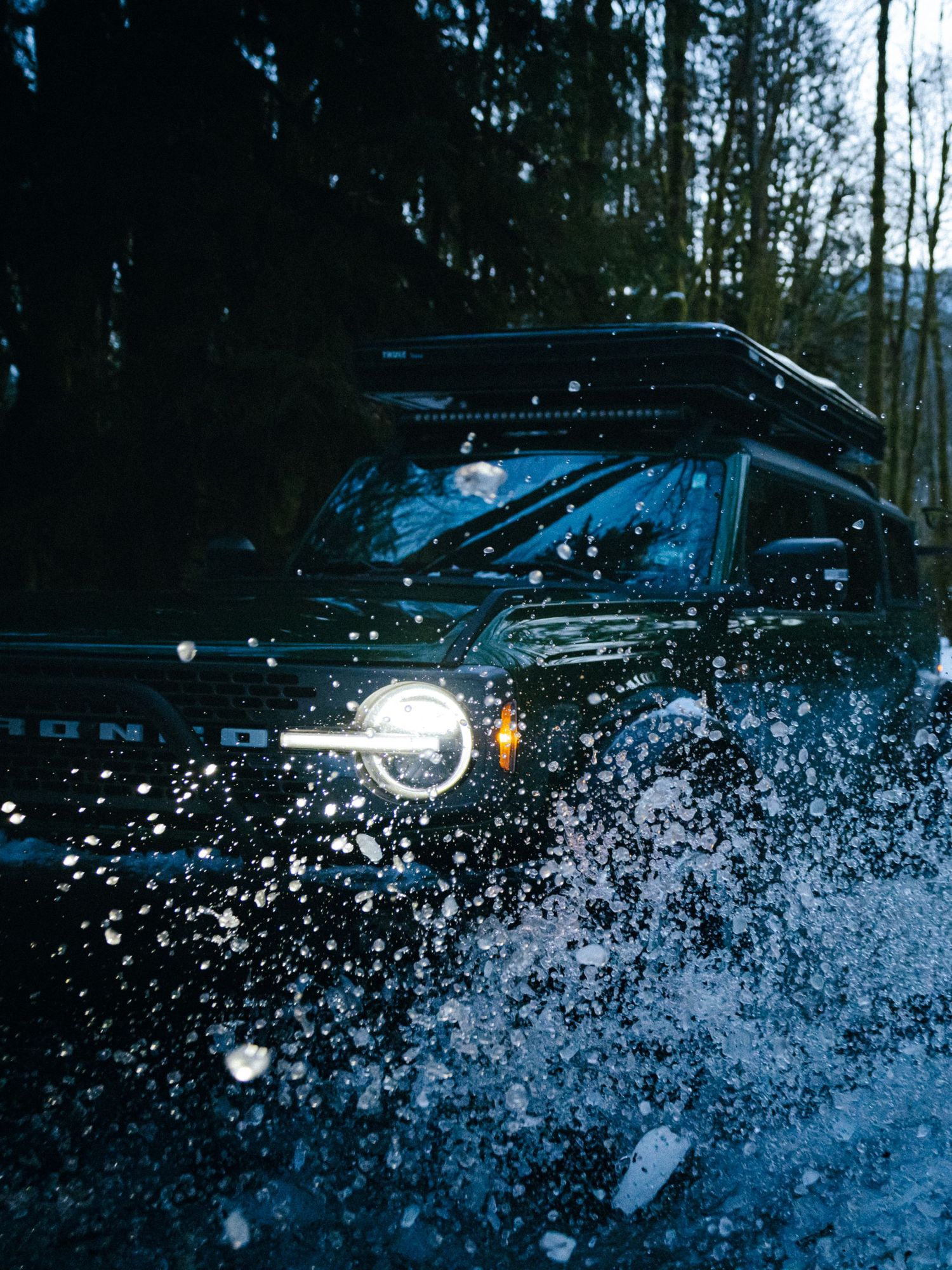 Ford Bronco splashing through water at night with headlights illuminating water spray in dark forest setting.