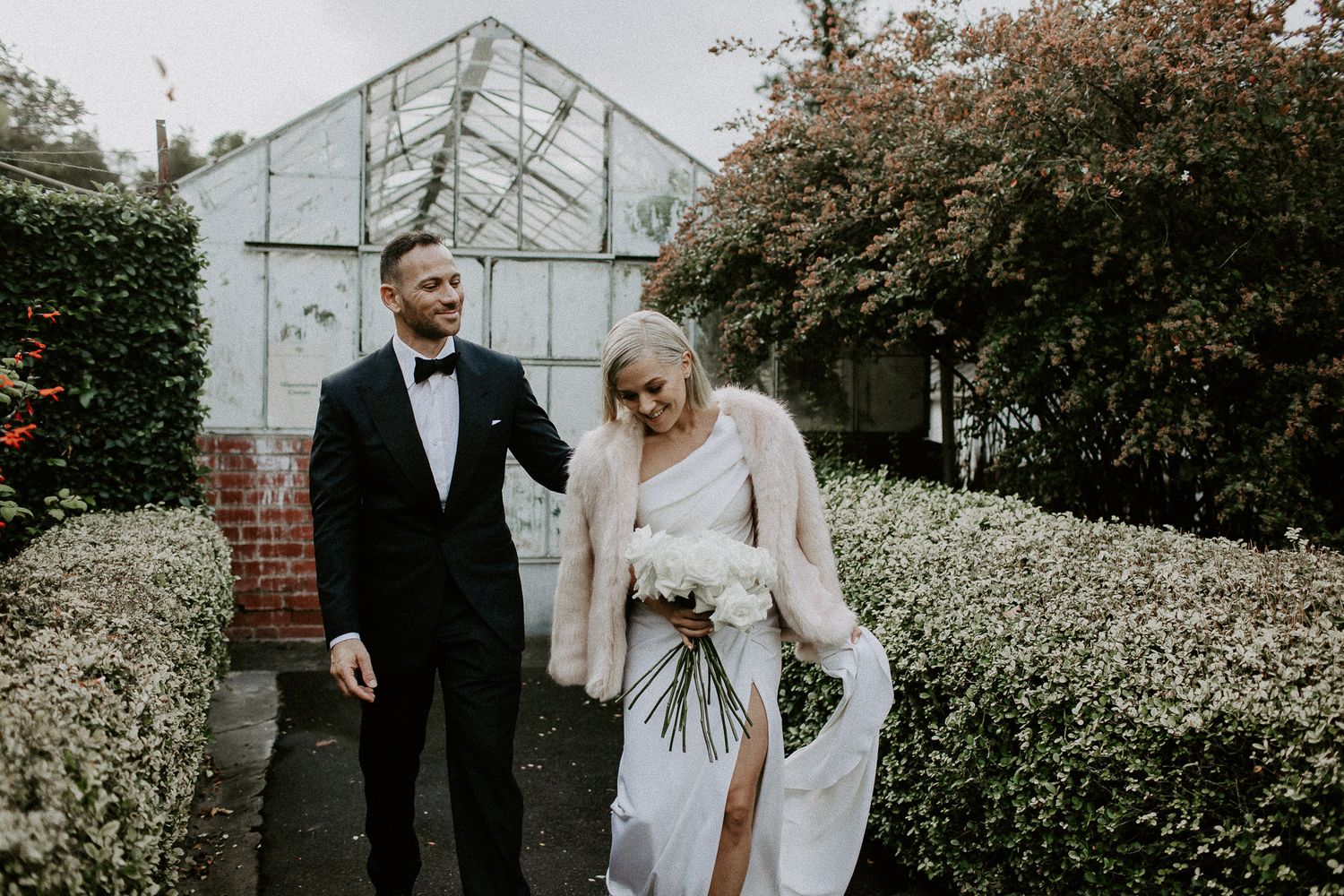 Couple in formal attire walks together near greenhouse in garden setting.