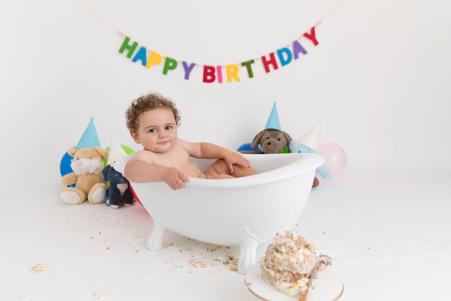 Baby in a white bathtub with stuffed animals, wearing a party hat, celebrating first birthday under a colorful Happy Birthday banner.