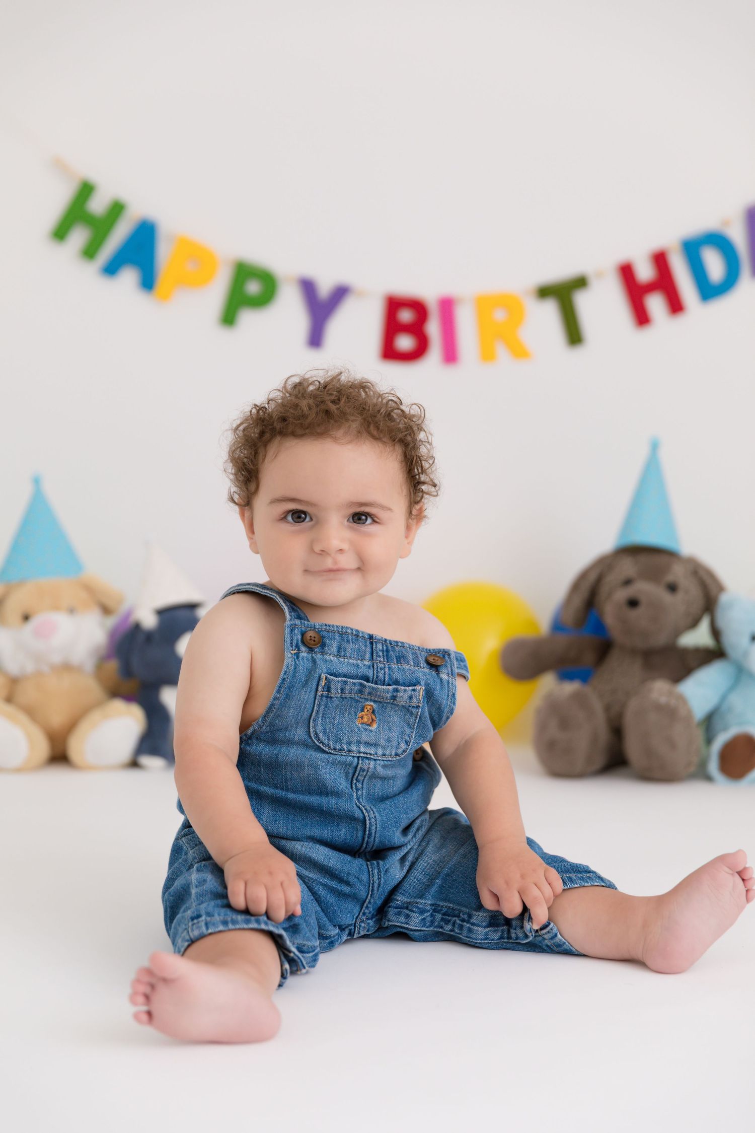 Cute baby in denim overalls sits barefoot in front of a Happy Birthday banner and stuffed animals.