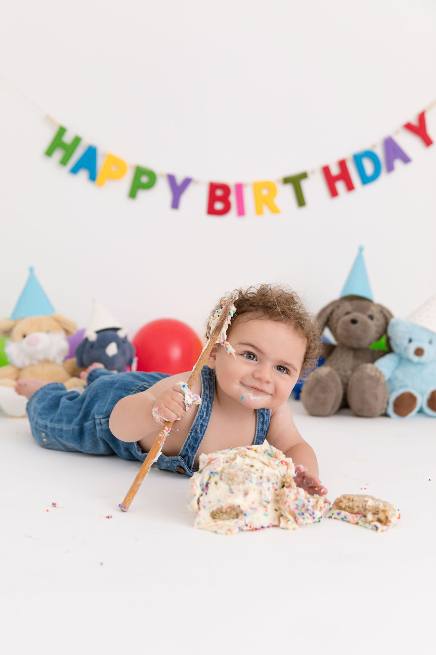 Baby in denim overalls smashing first birthday cake with a Happy Birthday banner and stuffed animals in background.