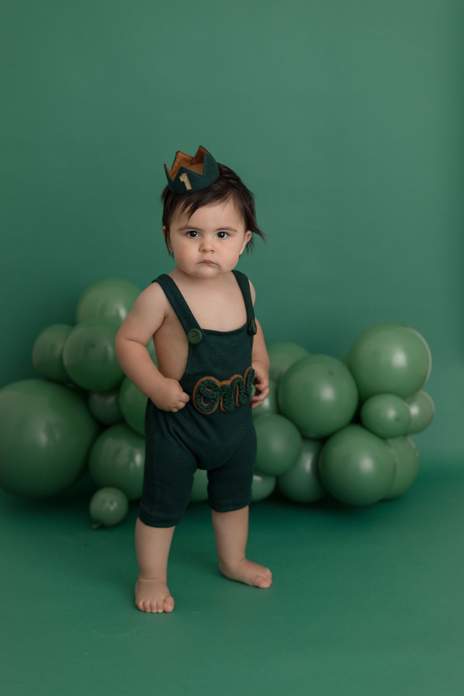 Baby in green overalls with 'one' embroidery stands before green balloons, wearing a crown for first birthday.