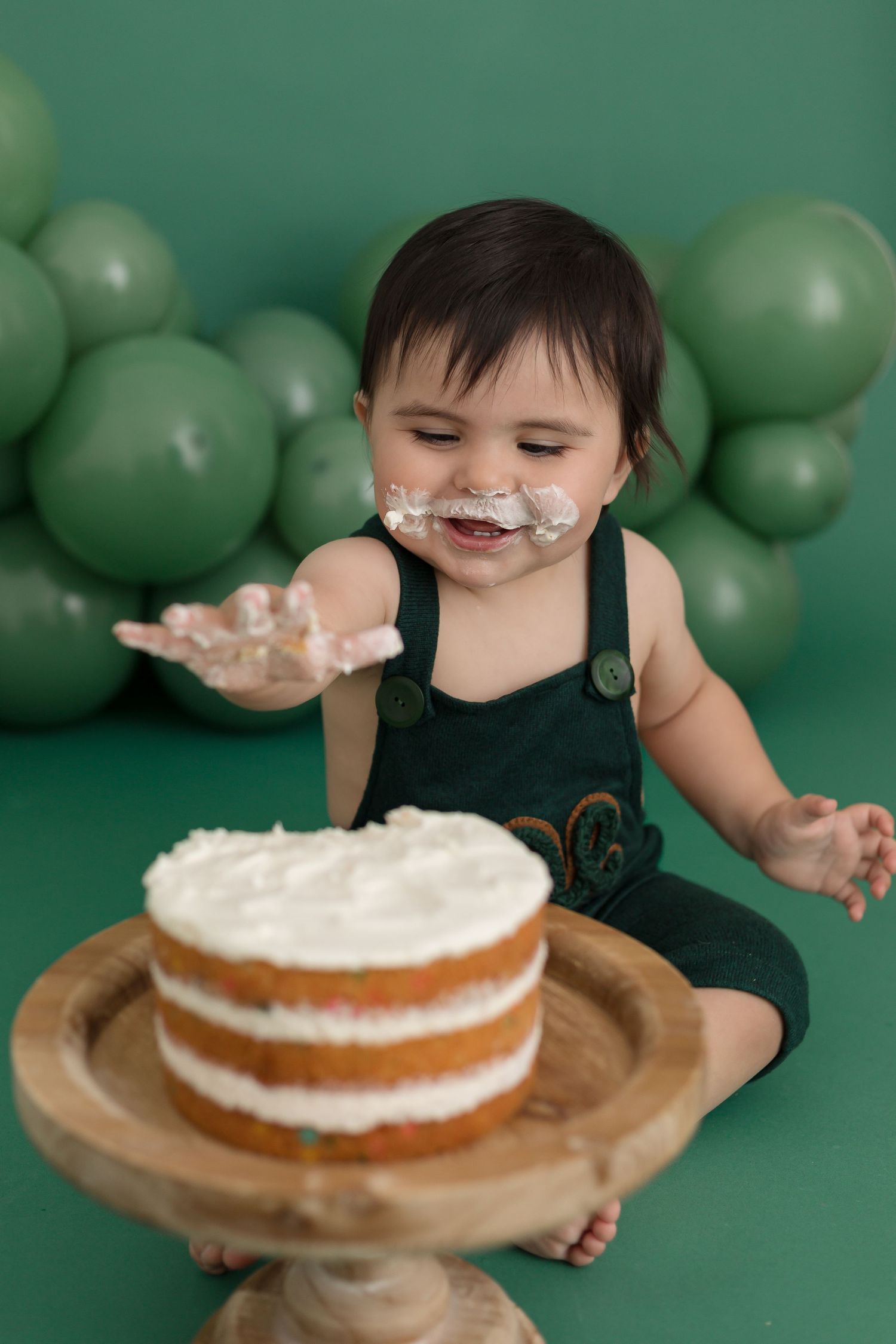 Smiling baby in green romper enjoys first birthday cake smash with white frosting on face, green balloon backdrop.