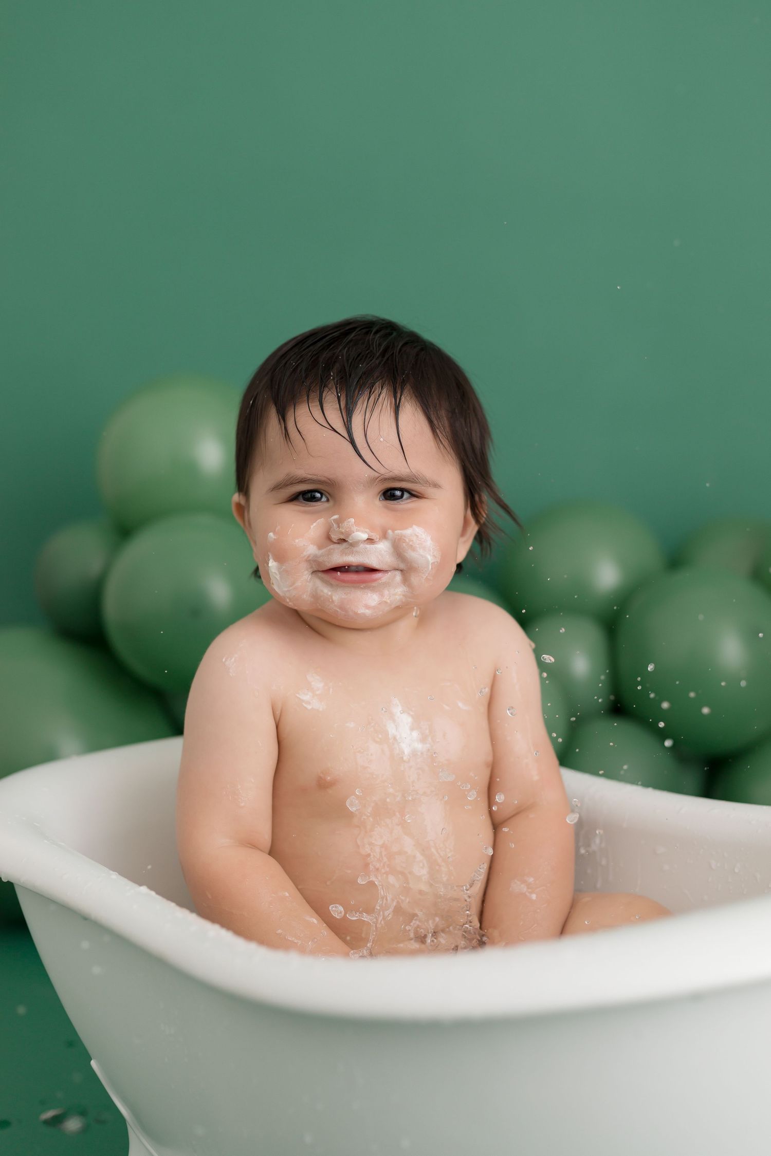 Happy baby sitting in a white bathtub with cake on face, green balloons backdrop, first birthday bath photo.