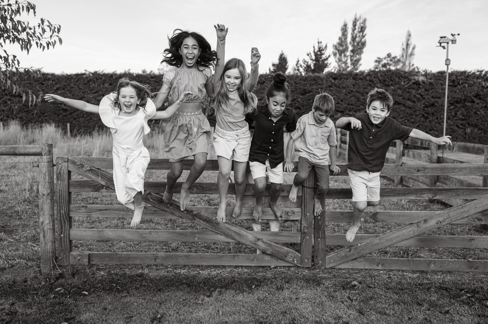 Black and white photo of children jumping together over a rustic wooden gate.