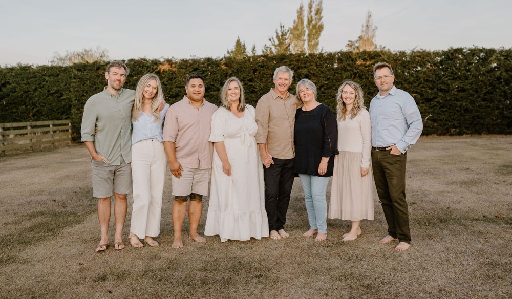 Family group in coordinated neutral outfits poses together outdoors against green hedge backdrop.