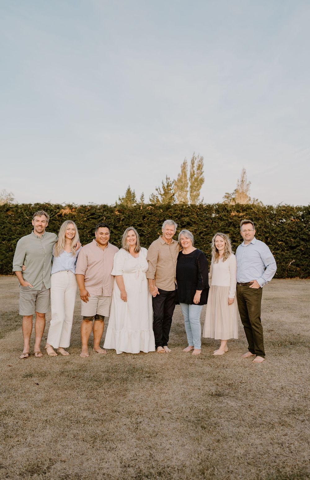 Extended family stands together on grass field with trimmed hedge backdrop during golden hour.