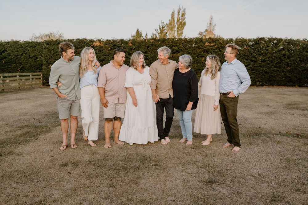 Warm-toned family photo of group standing together on dirt ground with manicured hedge background.