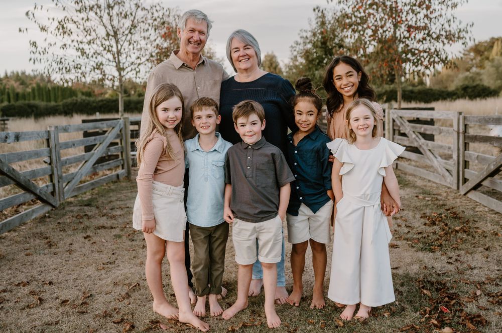 Multi-generational group poses together barefoot by wooden fence during sunset photoshoot.