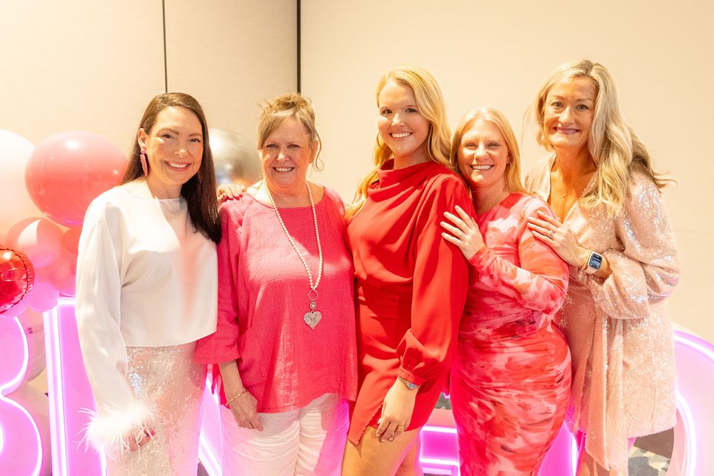 A group gathering with pink balloons and decorations wearing coordinated pink and white outfits at a celebration event.