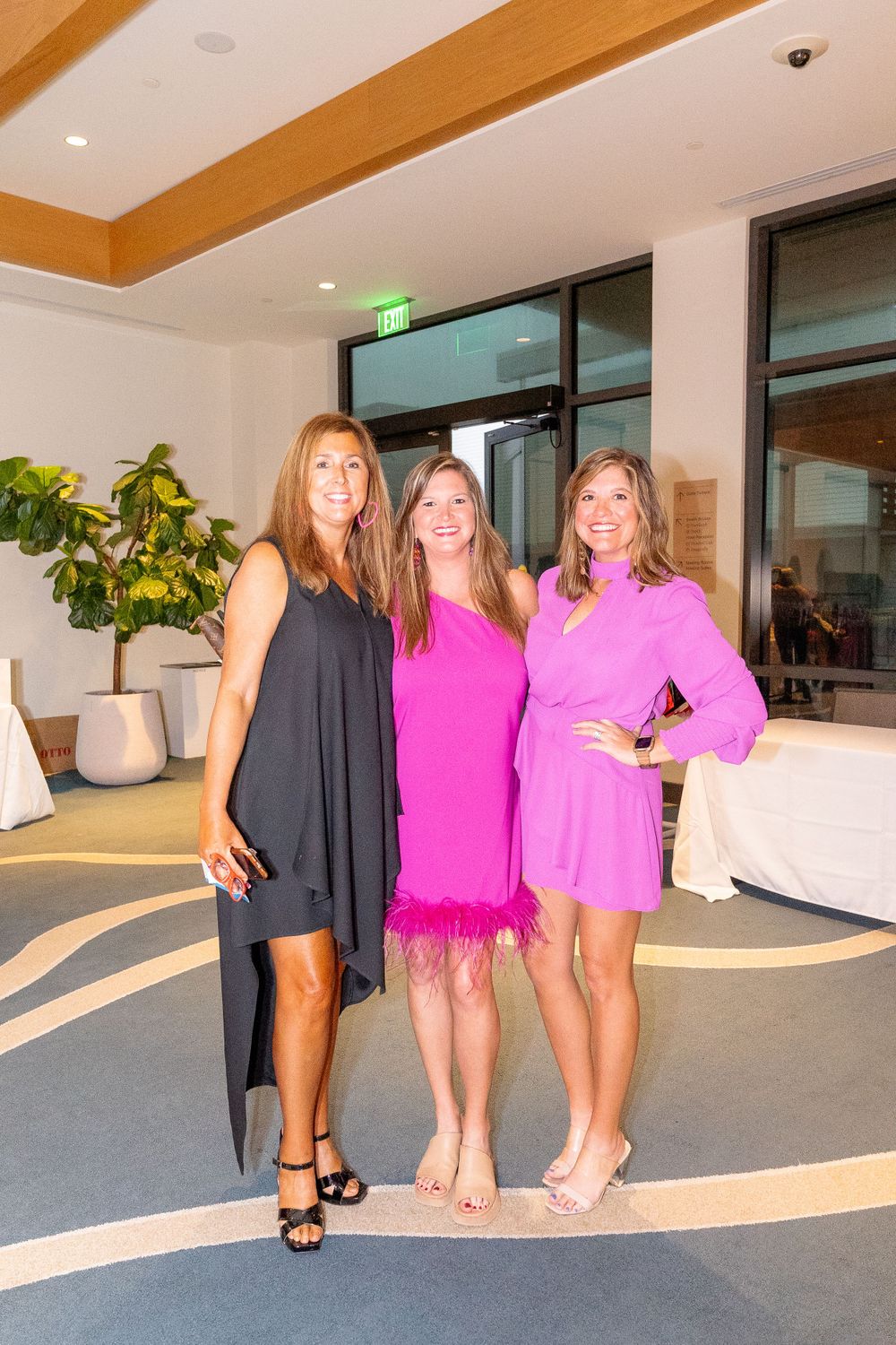 Three women pose together in a modern lobby wearing cocktail dresses in black and pink tones.