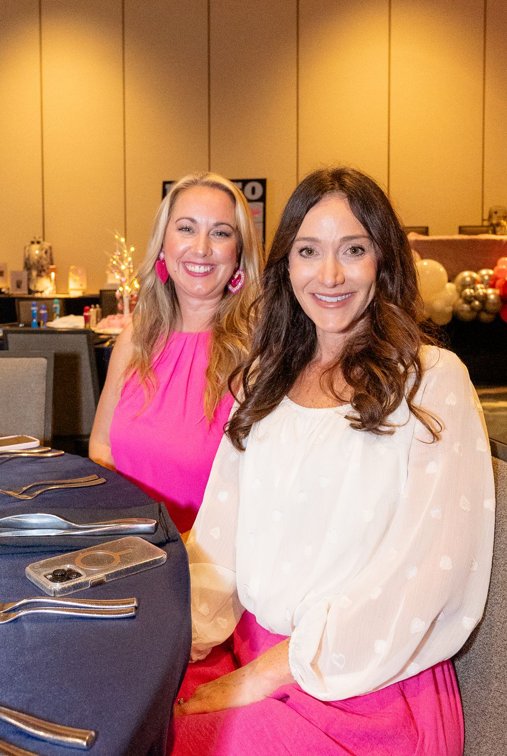Two women sitting at a formal dinner table setup with place settings and decorations.