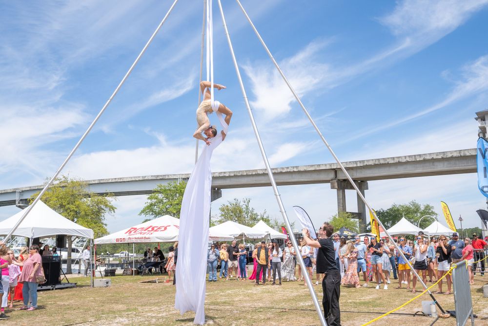 An aerial performer in white fabric performs under a tent at an outdoor festival with a bridge in background.