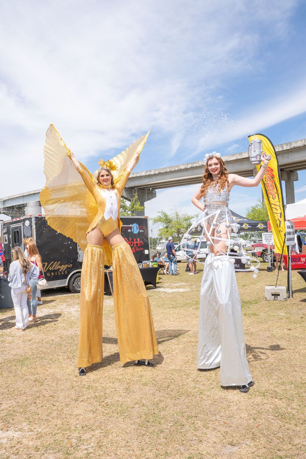 Performers on stilts wearing gold and white costumes entertain crowds at an outdoor festival.