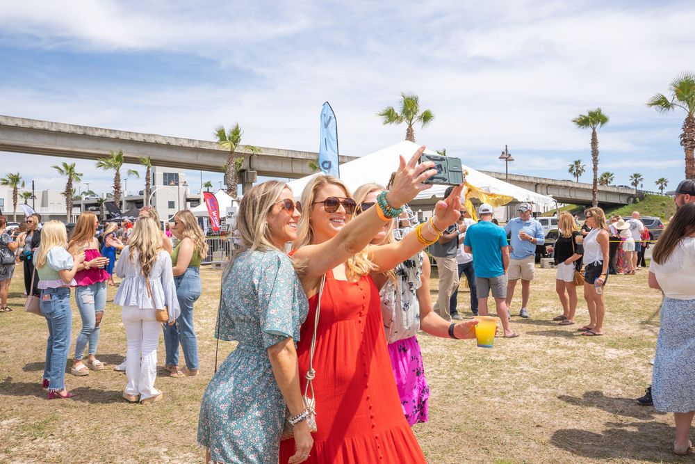 Guests taking selfies at an outdoor festival with palm trees and an overpass in the background.