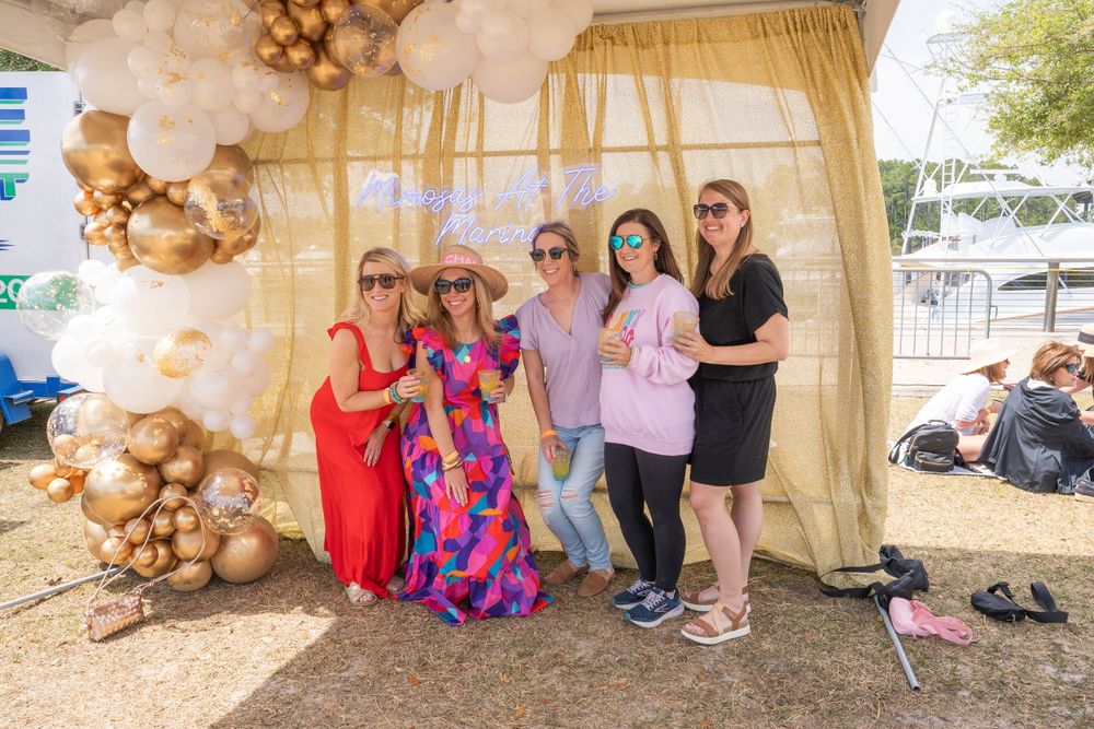 Attendees pose in a decorated gazebo with gold and white balloon arch at an outdoor celebration.