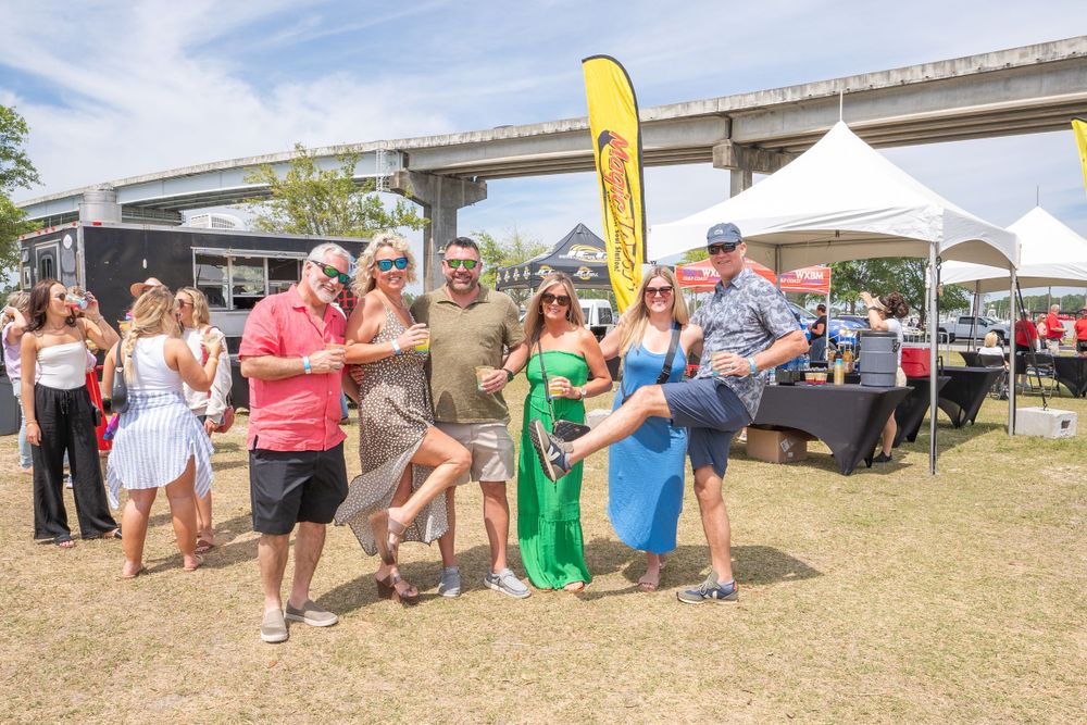A lively group posing together at an outdoor event with festival tents and banners visible.