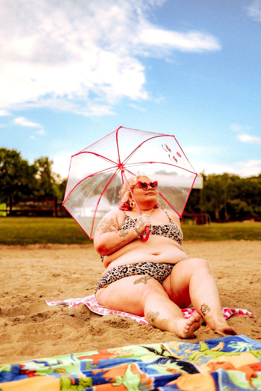 Person lounging under red umbrella on colorful beach blanket in sunny field.