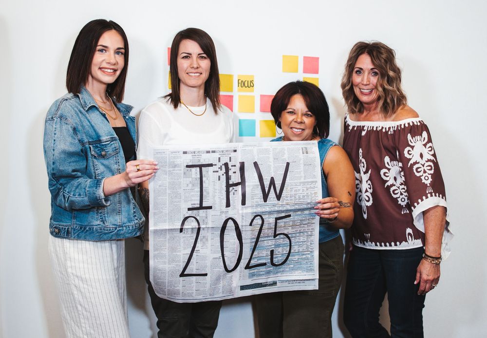 Group of coworkers hold a sign reading IHW 2025 while standing against a white background with sticky notes behind them.