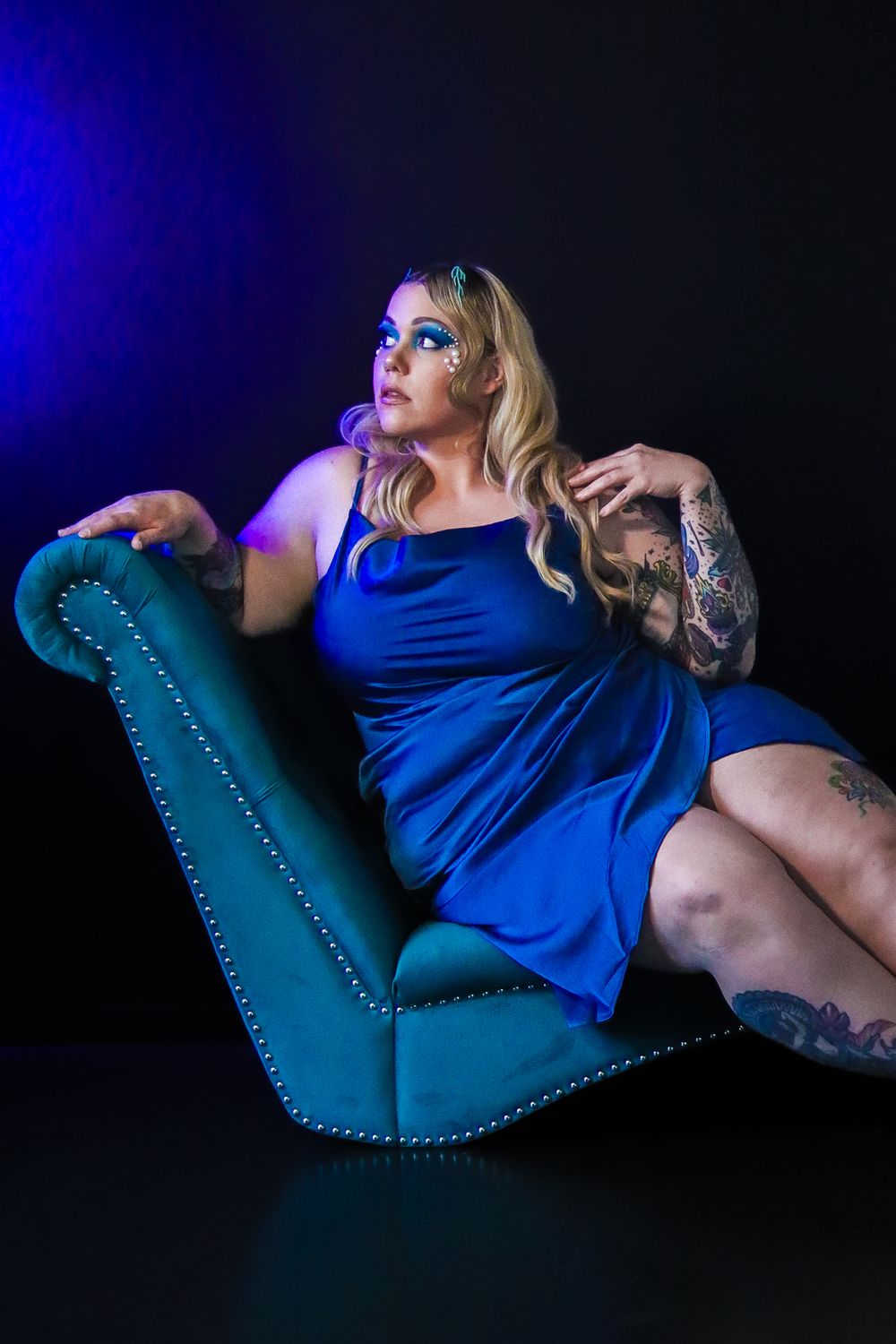 Artistic portrait on a teal chaise lounge featuring blue dress and tattoo sleeve against dramatic lighting.