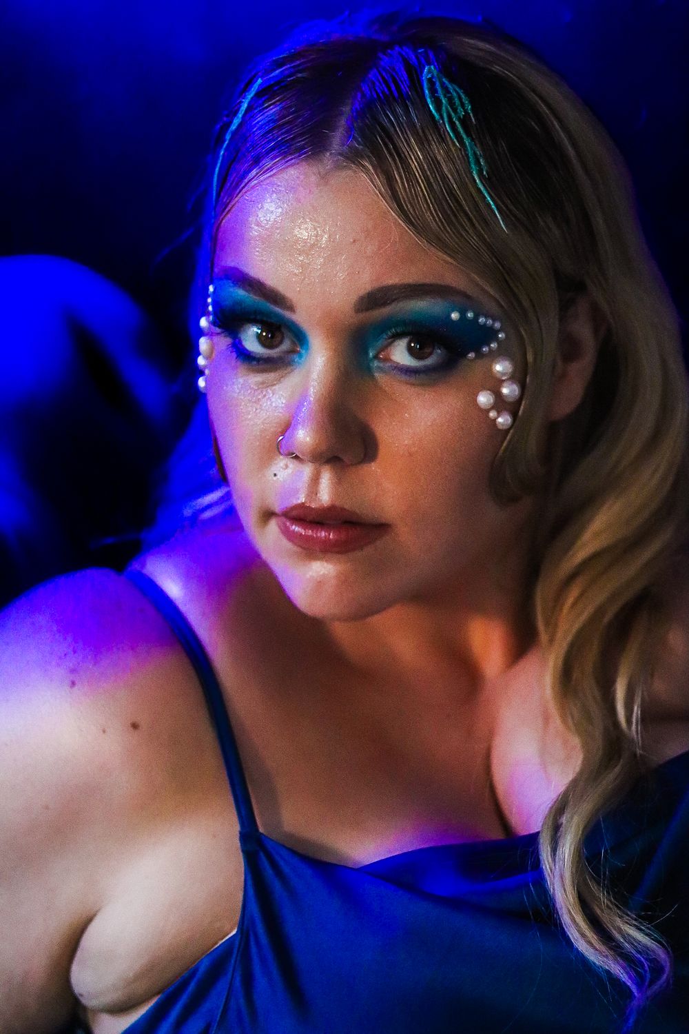 Close-up portrait with blue lighting showing dramatic eye makeup with pearl details and wavy blonde hair.