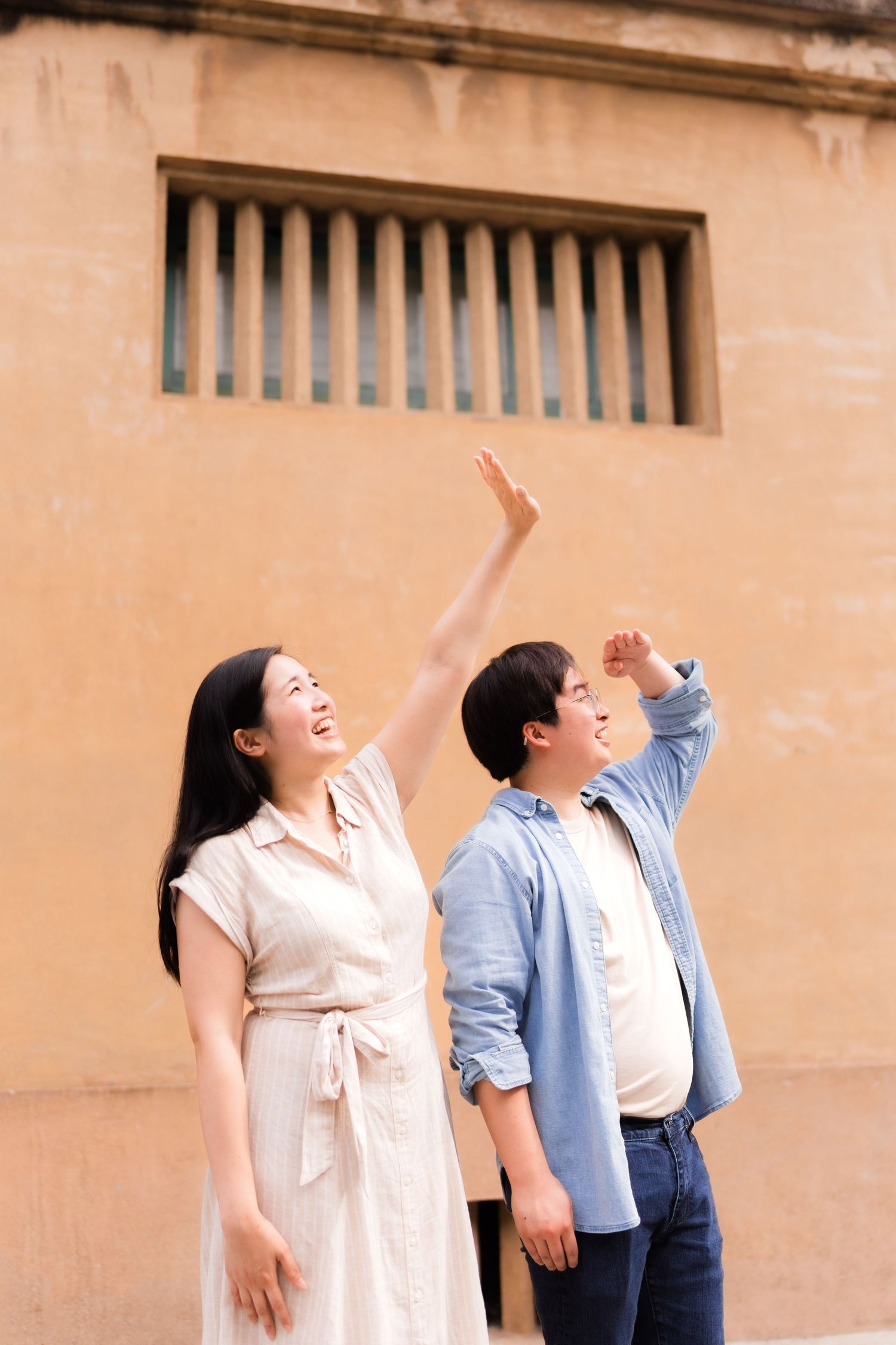 Two people gesture upward while standing against a beige wall with barred windows.
