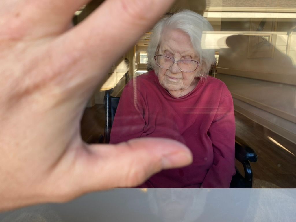 Large hand presses against glass windows framing an elderly women sitting inside who's sleeping in a wheel chair