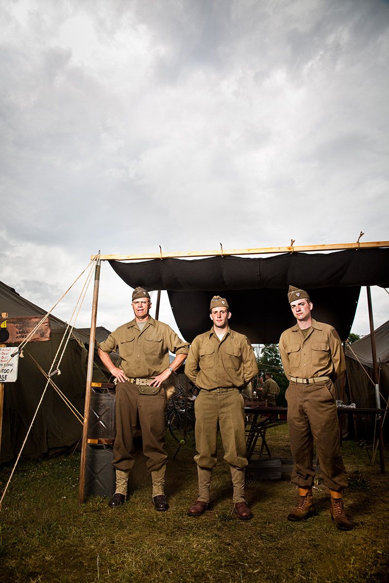 Three men in WWII military uniforms standing before army tent under cloudy sky.