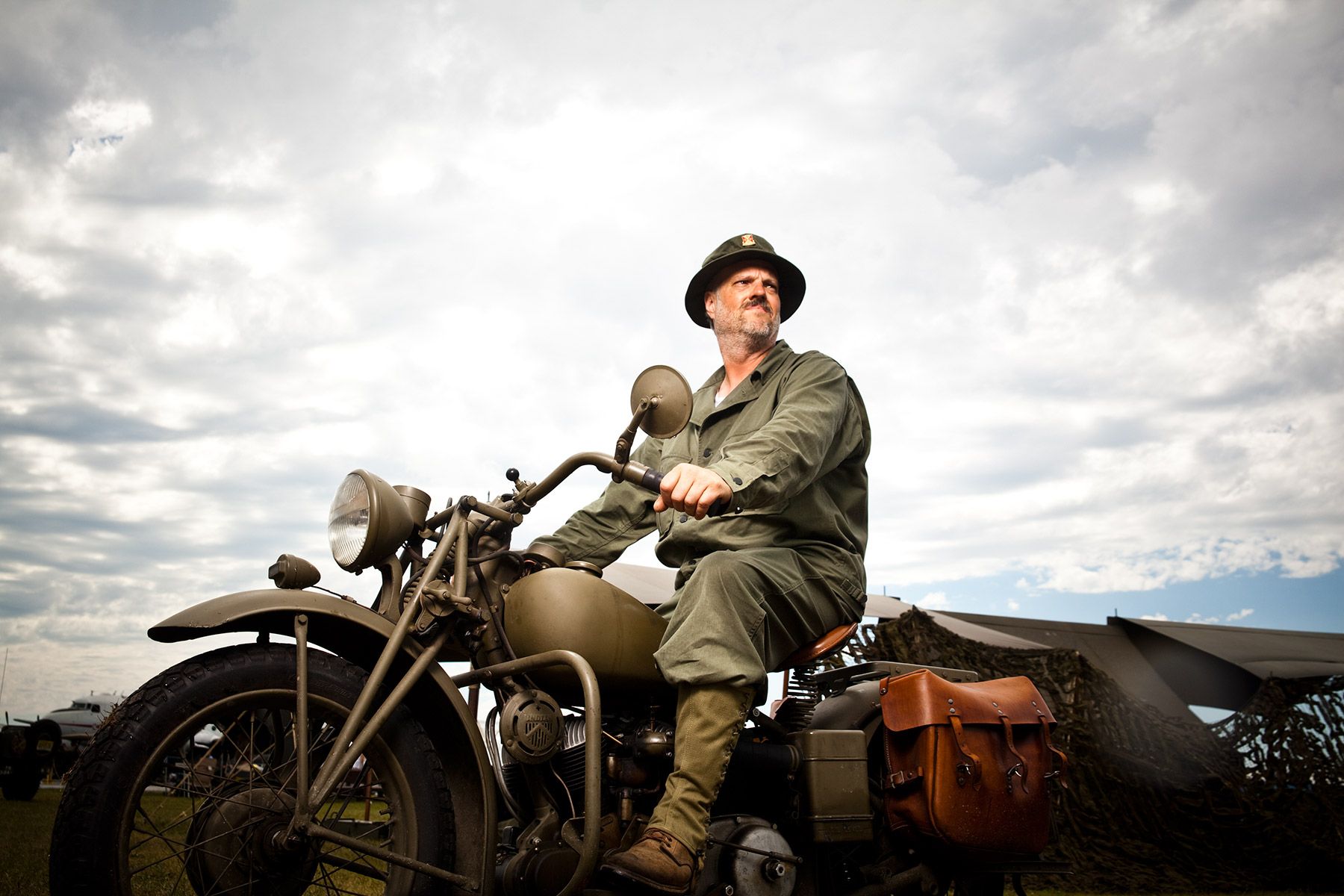 Man in military uniform sitting on vintage WWII motorcycle under dramatic cloudy sky.