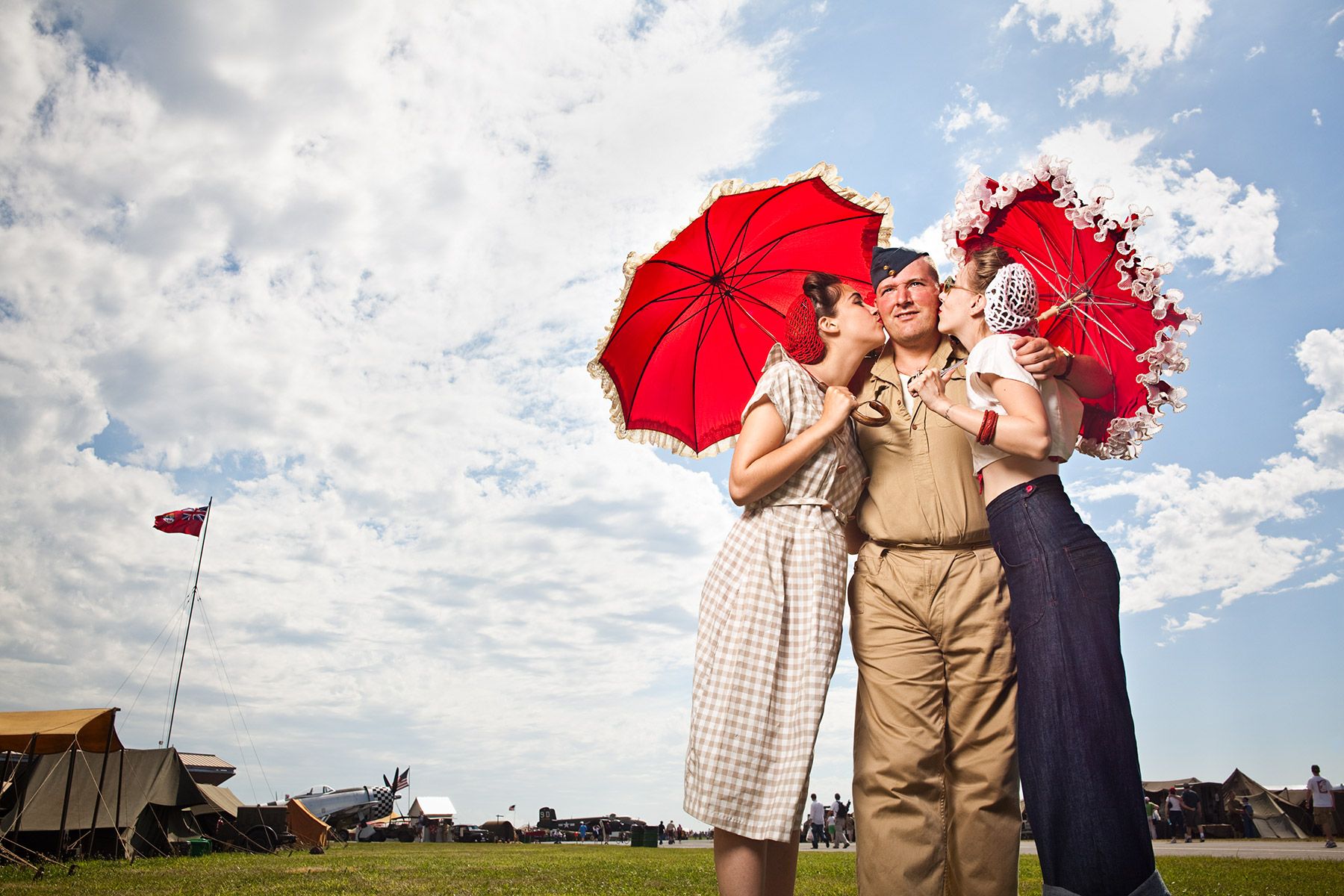 Three people in 1940s attire holding red umbrellas on airfield under blue sky.