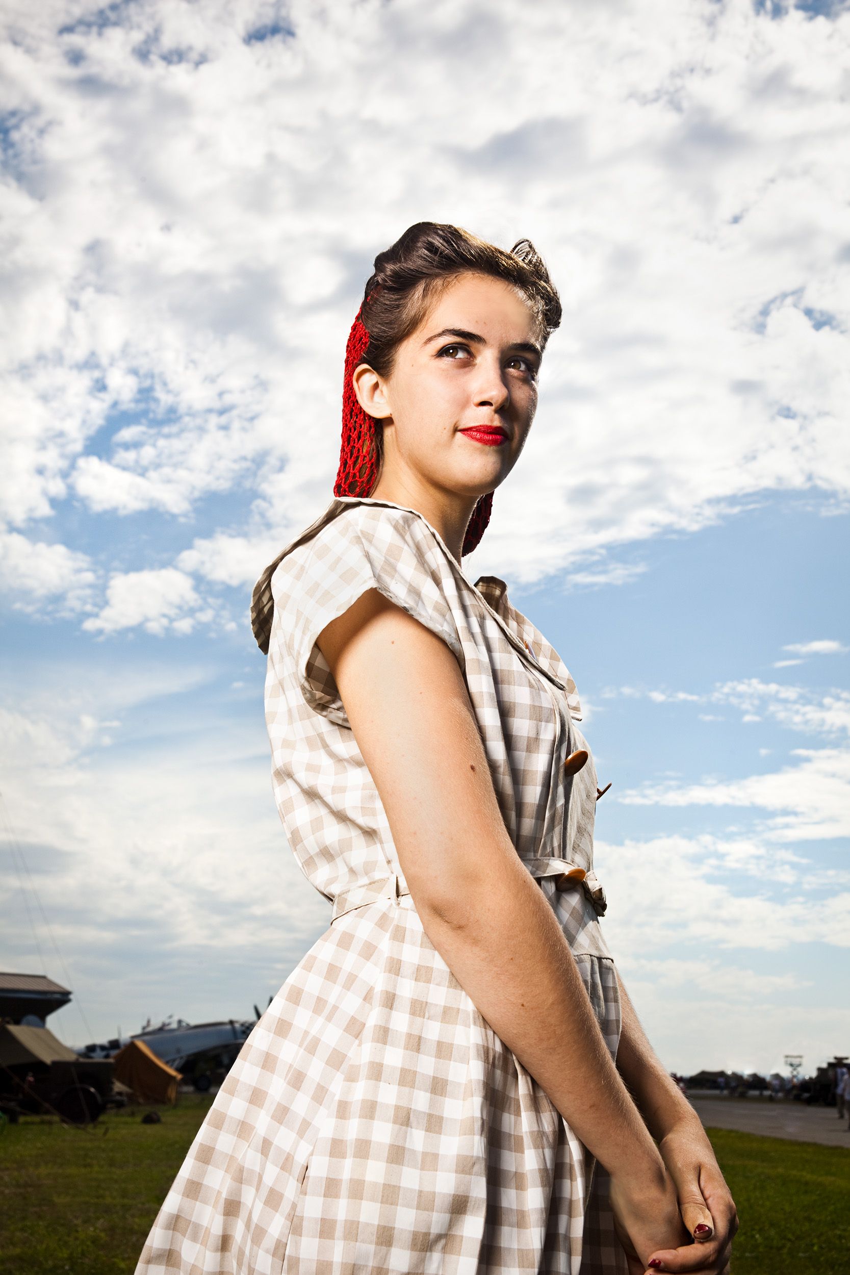 Young woman in vintage 1950s plaid dress with red hair accessory posing under cloudy sky.
