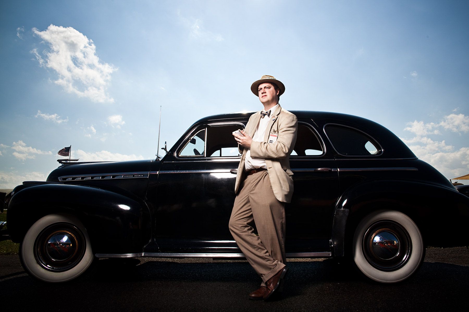 Man in vintage suit leaning against classic black 1940s car under blue sky.