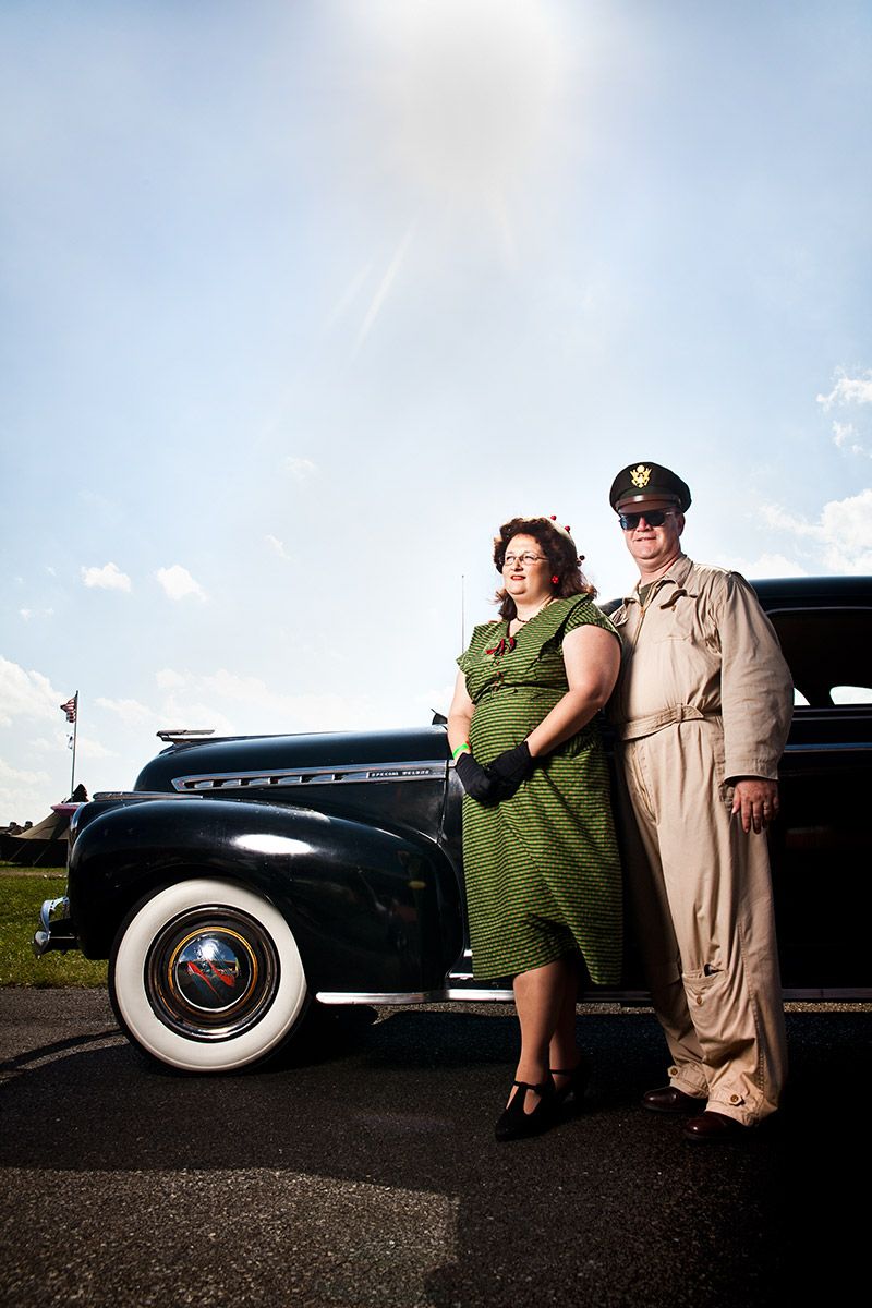 Couple in 1940s vintage attire posing beside classic black car outdoors.