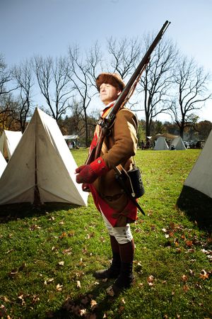 Revolutionary War reenactor in period costume carrying musket at outdoor encampment.