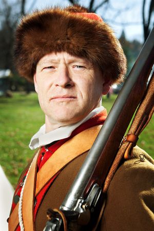 Man in fur hat and frontier costume holding musket rifle outdoors.